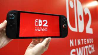 Hands hold a Nintendo Switch 2 console near a display with the logo on it, at an event. | Fumiyasu Nakatsuji/Jiji Press/Newscom
