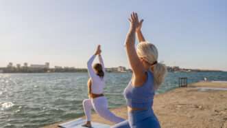 Two women in yoga poses on the shoreline of a beach. | Msfiolent | Dreamstime.com